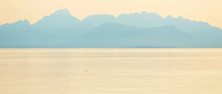 Pale layered mountains fading into mist across a calm sea at dusk