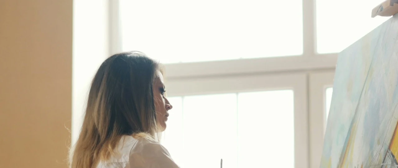 A woman seen from behind in a sunlit studio, mixing paint on a palette in front of a tall window