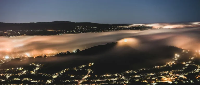 A city seen from a hillside at night with fog rolling through the valley under a starry sky