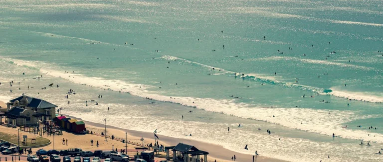 Dozens of surfers in the waves off Muizenberg beach, Cape Town, on a bright day, seen from above