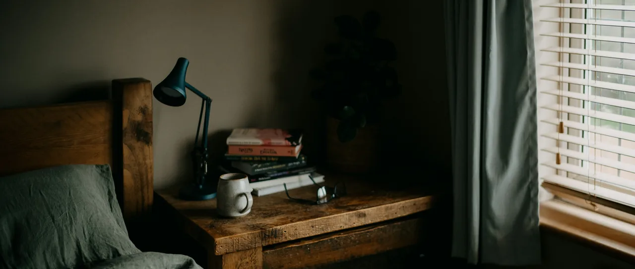 A dim bedroom at dusk, a blue reading lamp lit on a wooden nightstand stacked with books, glasses and a mug