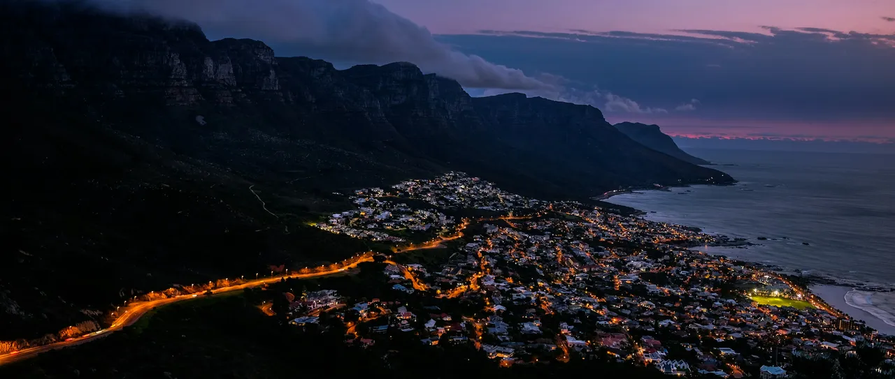 Aerial view of a coastal suburb lit up at night below Lion's Head, with a pink dusk sky, Cape Town