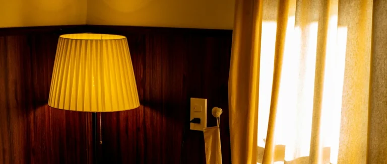 A pleated lamp glowing beside sheer curtains at dusk