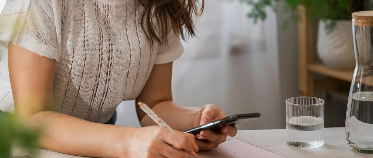 Hands writing notes while checking a phone at a bright kitchen table