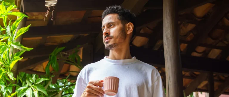 A man holding a pink coffee cup looks out from a wooden veranda surrounded by green leaves