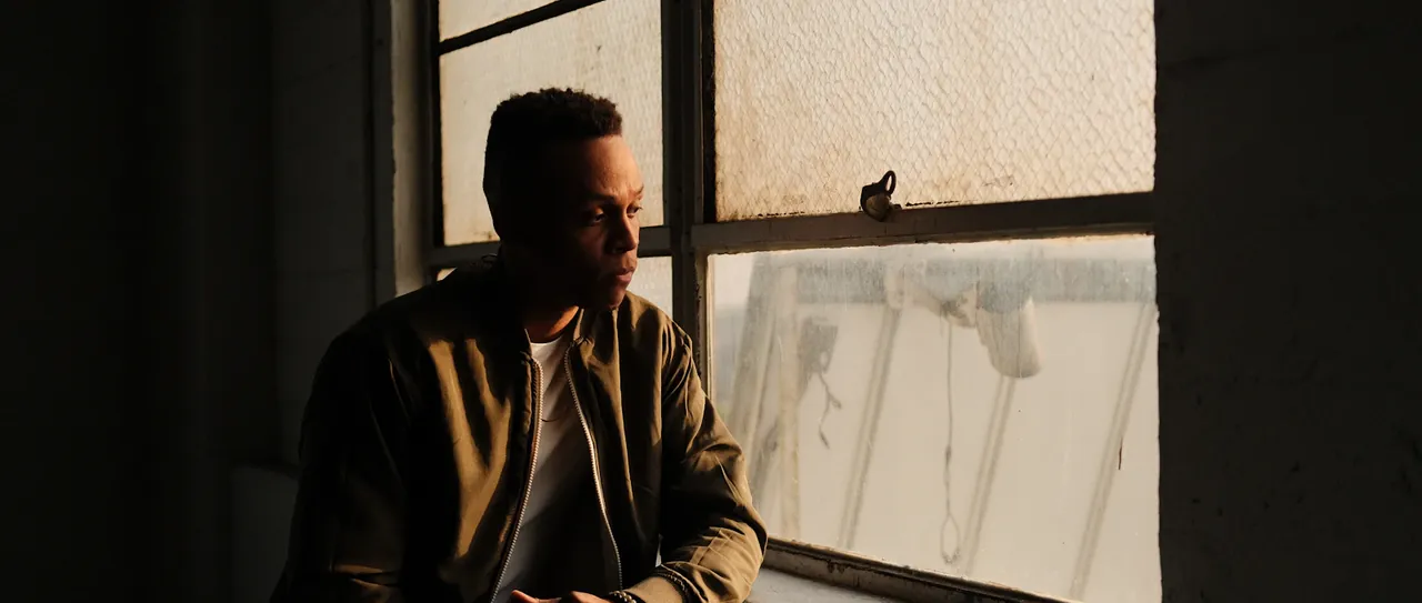 A man sits by a large window in warm evening light, looking down in thought