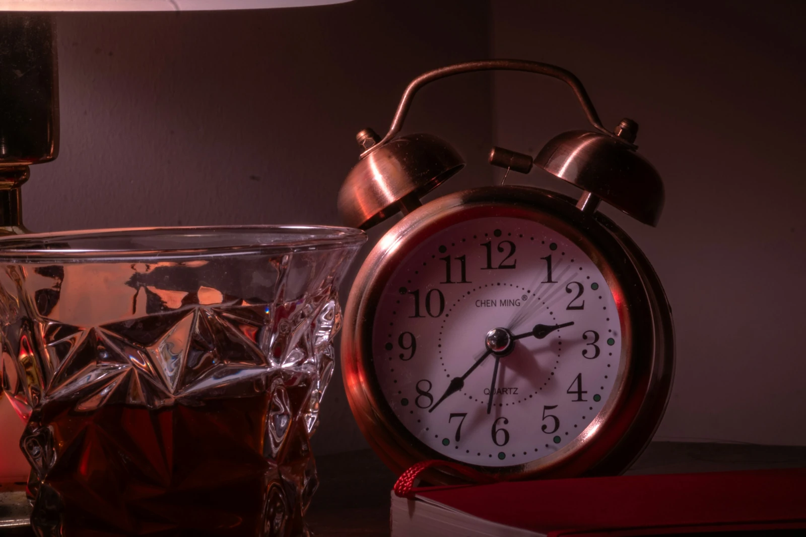 An old-style alarm clock standing beside a cut-glass tumbler of amber drink on a dark nightstand