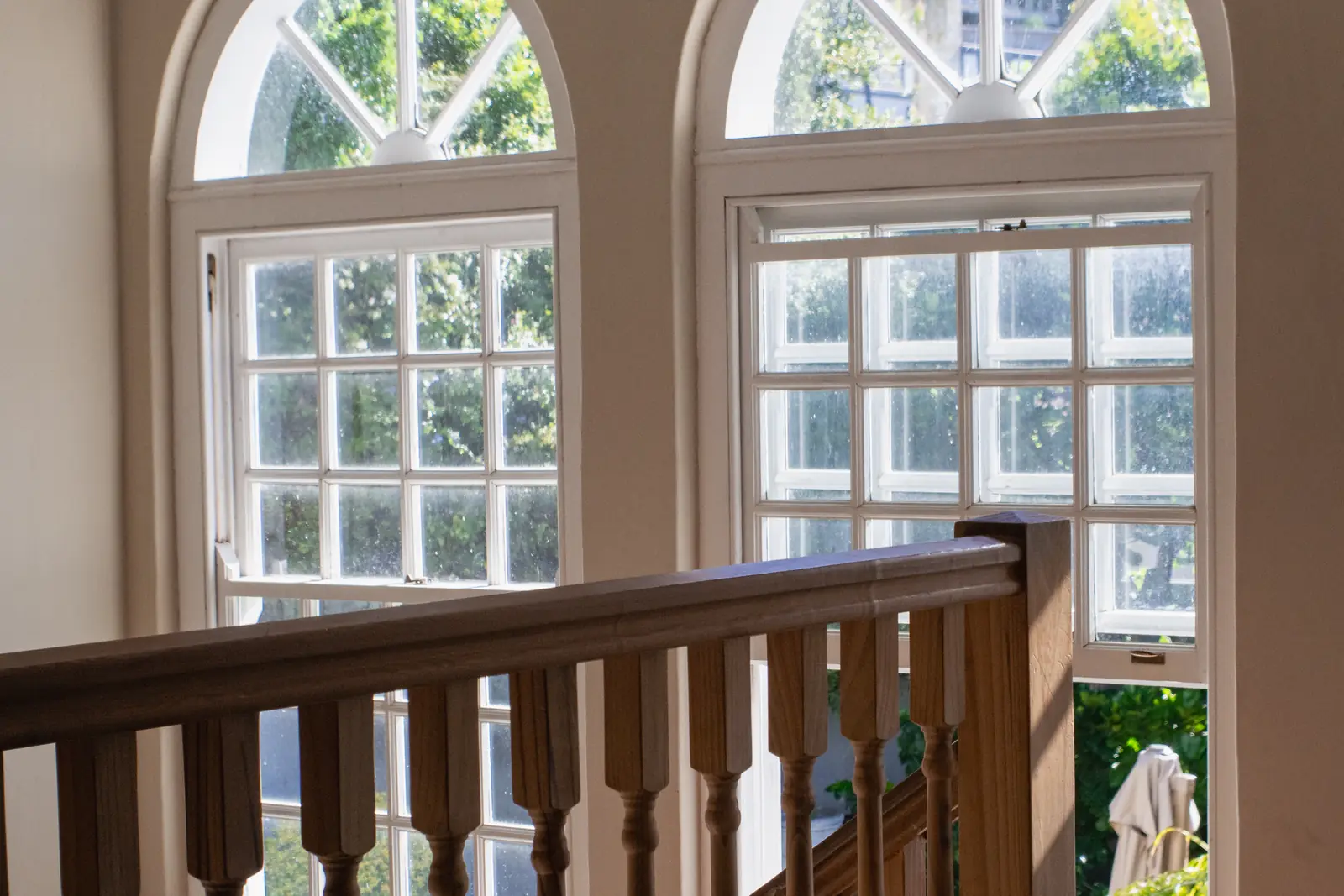 Two tall arched sash windows above a wooden staircase banister, with daylight and garden greenery outside