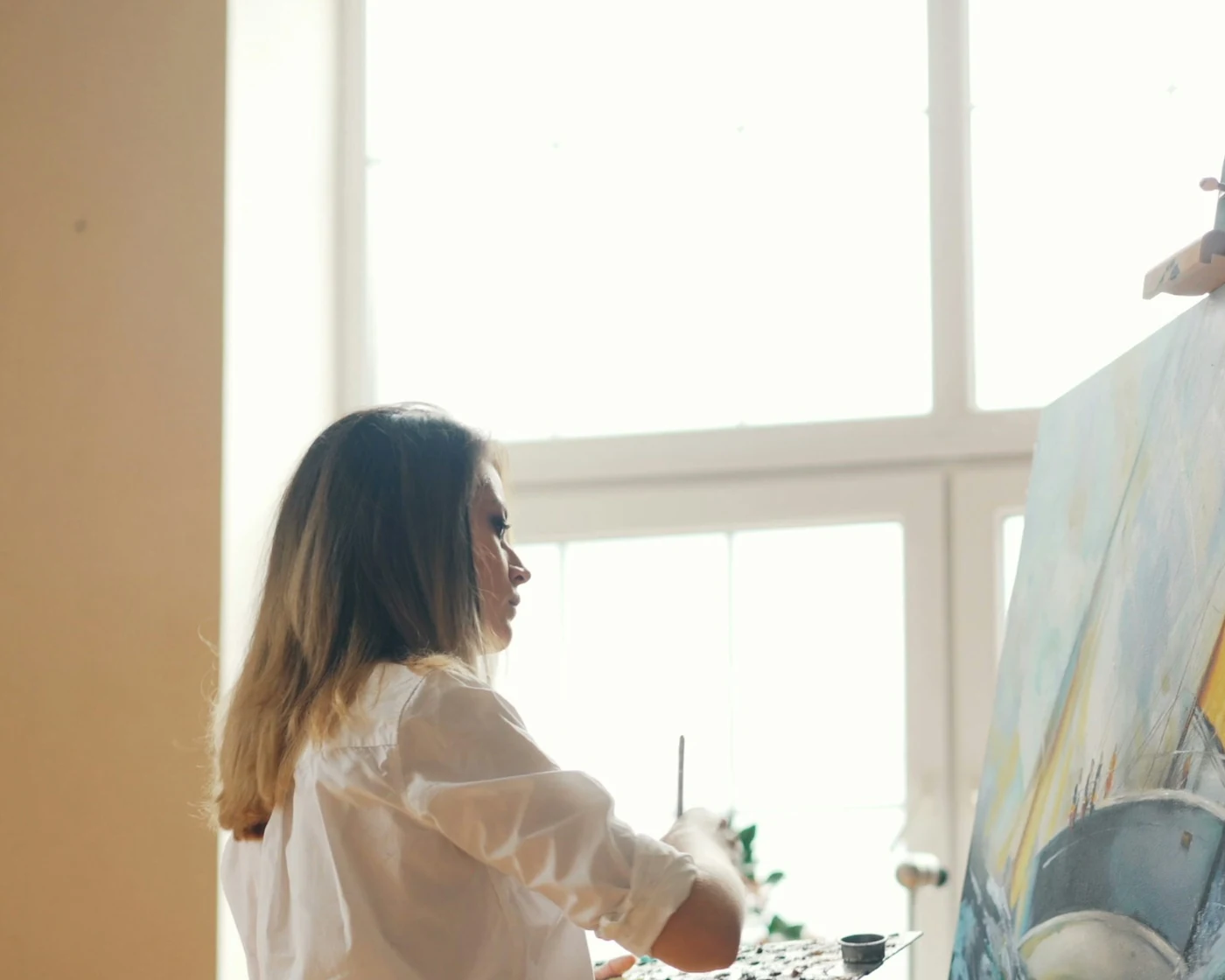A woman seen from behind in a sunlit studio, mixing paint on a palette as she works at an easel in front of a tall window
