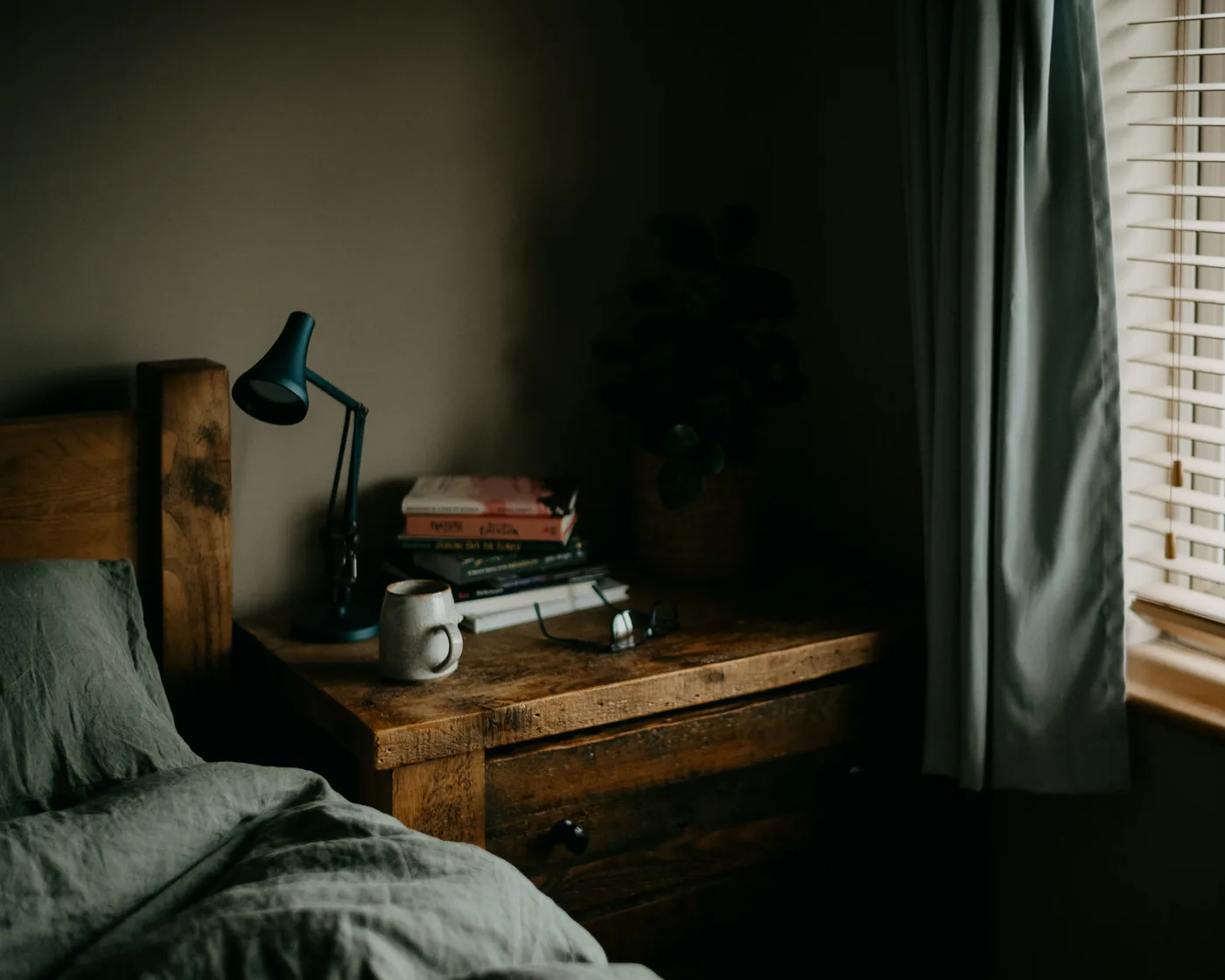 A dim bedroom at dusk, a blue reading lamp lit on a wooden nightstand stacked with books, glasses and a mug