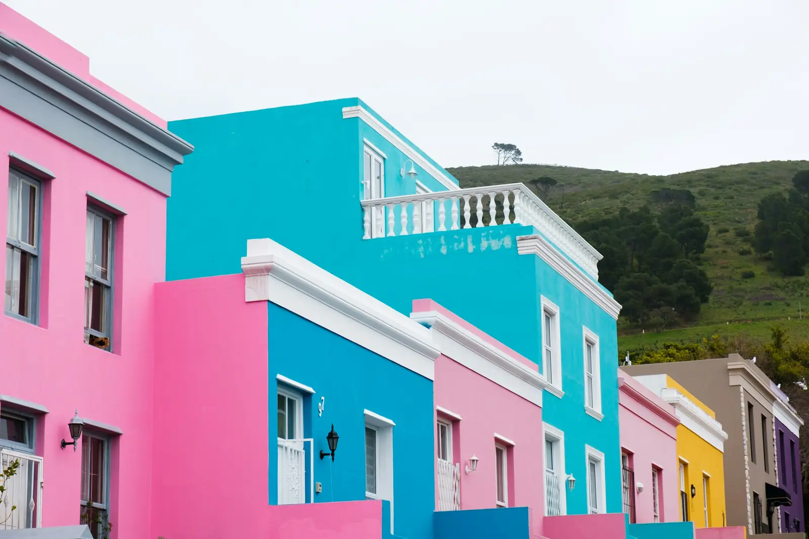 Pink, blue and yellow row houses in Bo-Kaap, Cape Town, below a green hillside