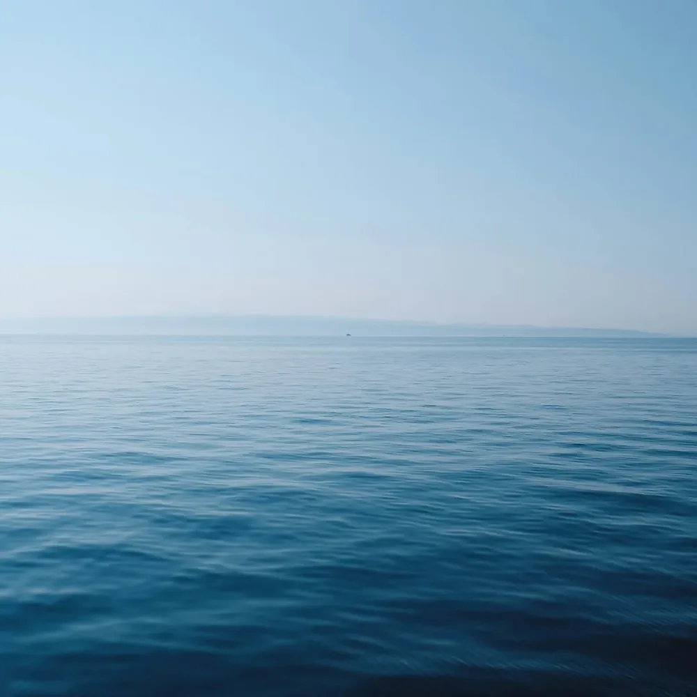 A calm, clear sea meeting a pale sky at the horizon, with faint hills in the distance