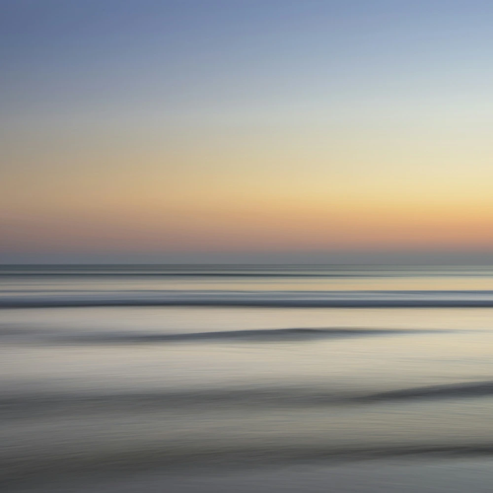 Calm sea at dawn, soft long-exposure water and a warm orange-to-blue sky at the horizon