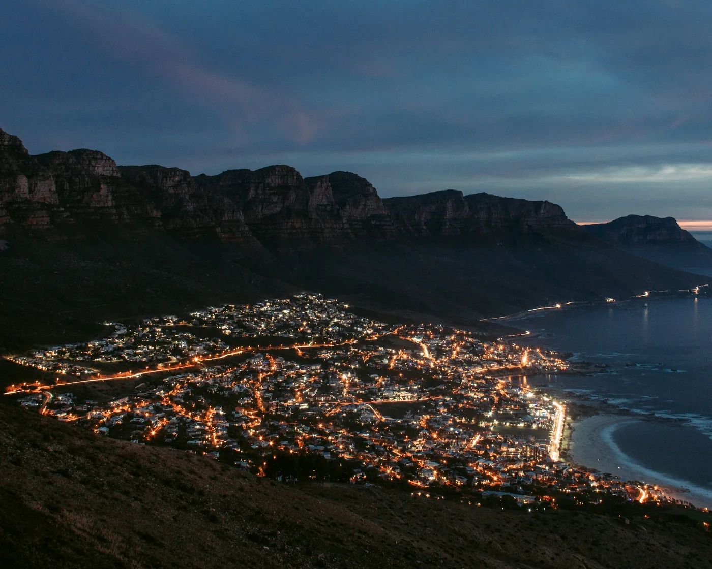 Camps Bay and the Atlantic seaboard lit up at dusk, seen from the mountainside above, sea and a rocky headland on the right