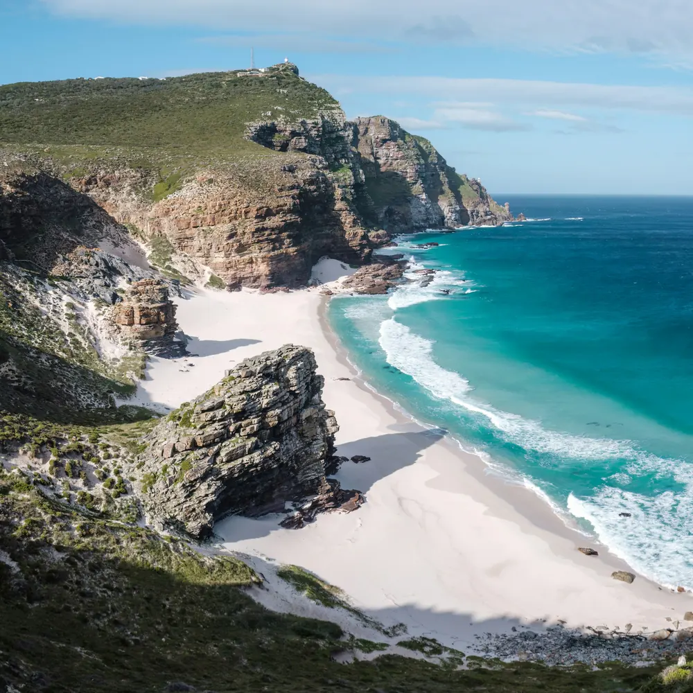 A white sand beach with turquoise waves below rocky cliffs at the Cape of Good Hope in bright daylight