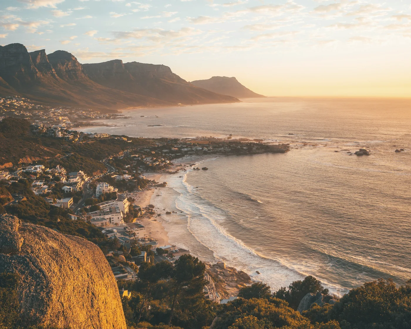 The Atlantic seaboard coastline in warm morning light, with granite boulders in the foreground and mountains along the shore, Cape Town