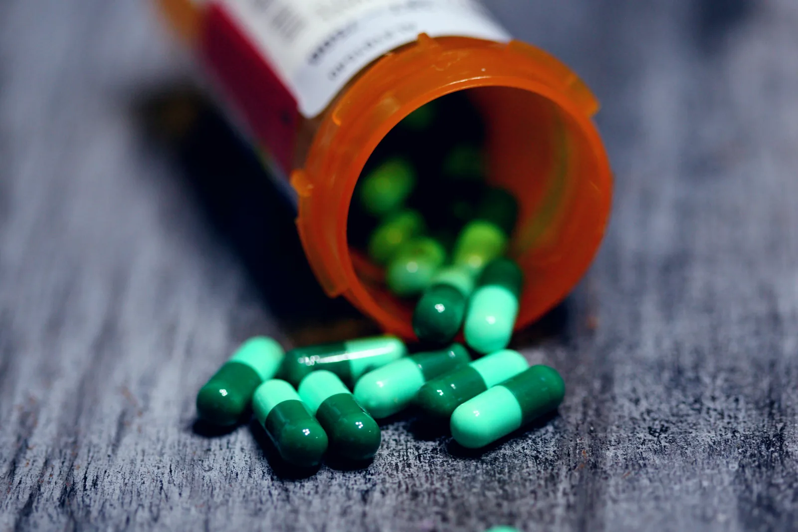 Green and white capsules spilling from a tipped-over orange pill bottle onto a wooden table, label out of focus