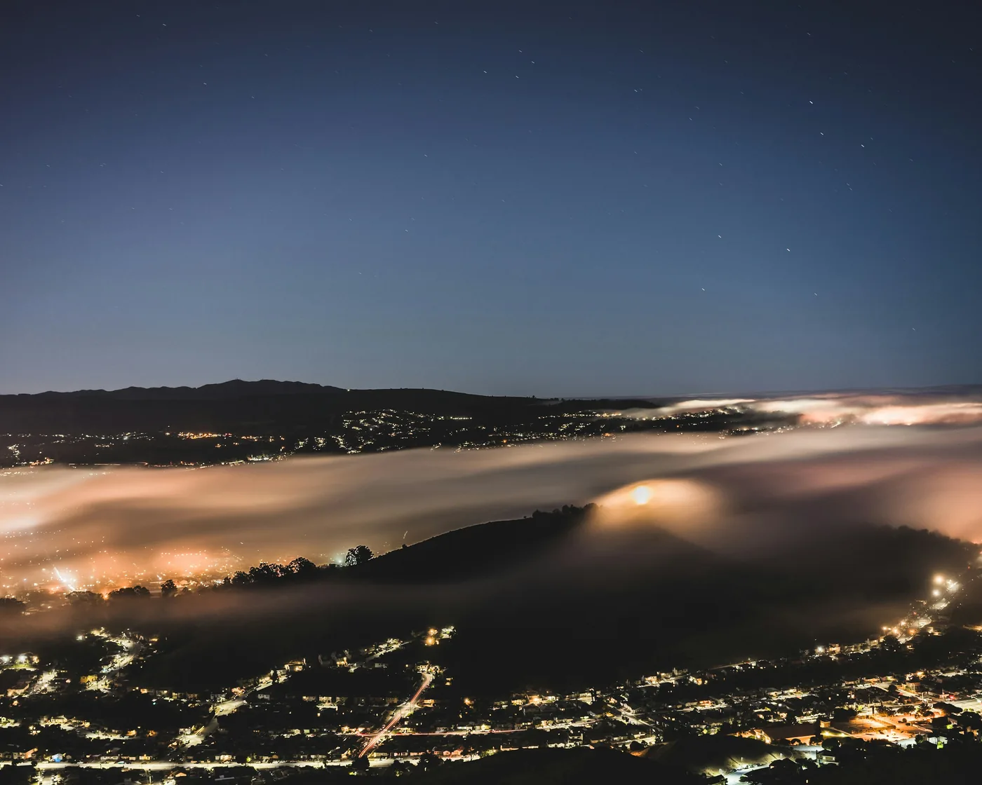 A city seen from a hillside at night, low fog rolling through the valley and catching the glow of streetlights under a starry sky
