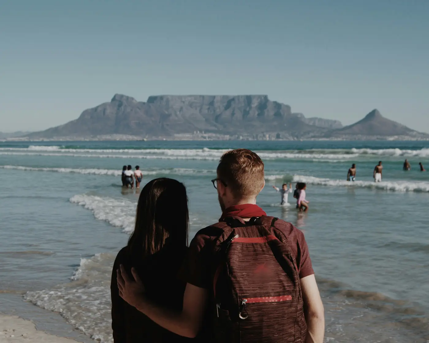 A couple stand arm in arm on the beach at Bloubergstrand looking across the bay at Table Mountain in Cape Town