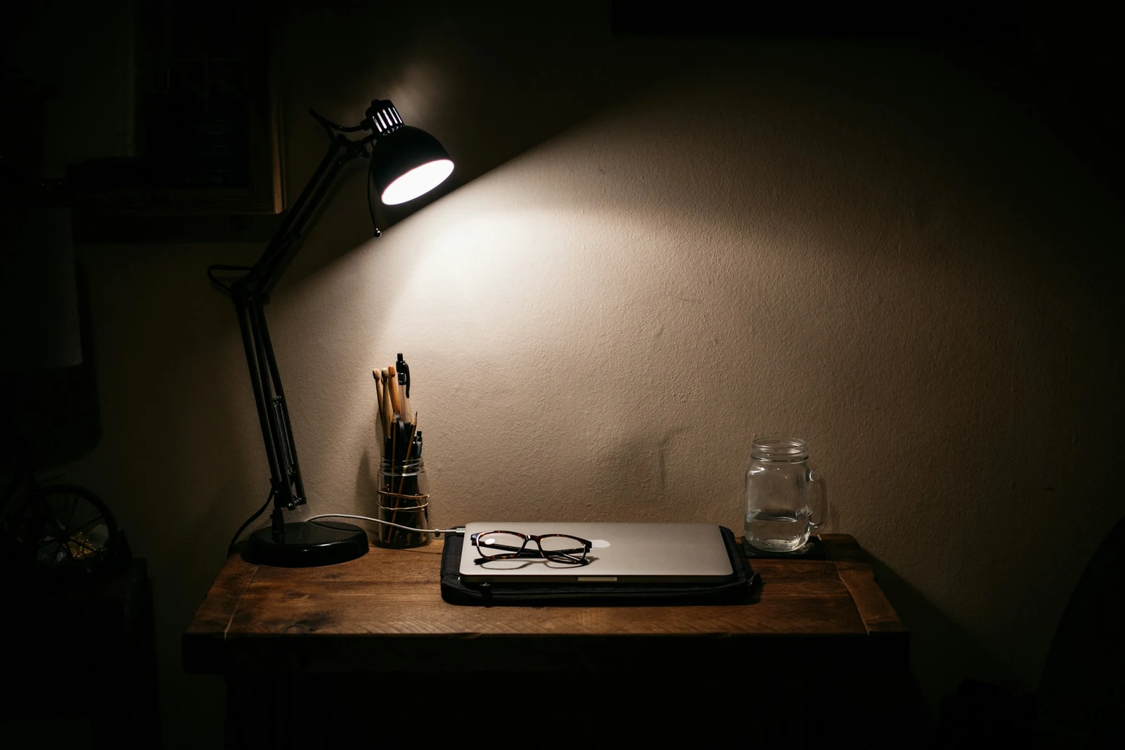 A lit desk lamp over glasses resting on a closed laptop and a jar of water at night