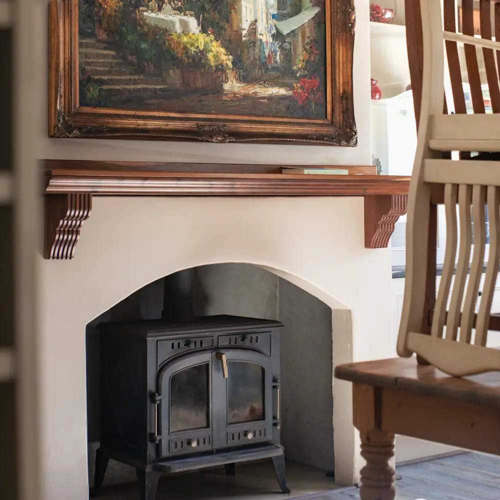 A wood burning stove in a white fireplace under a gilt framed oil painting, with a wooden chair beside it