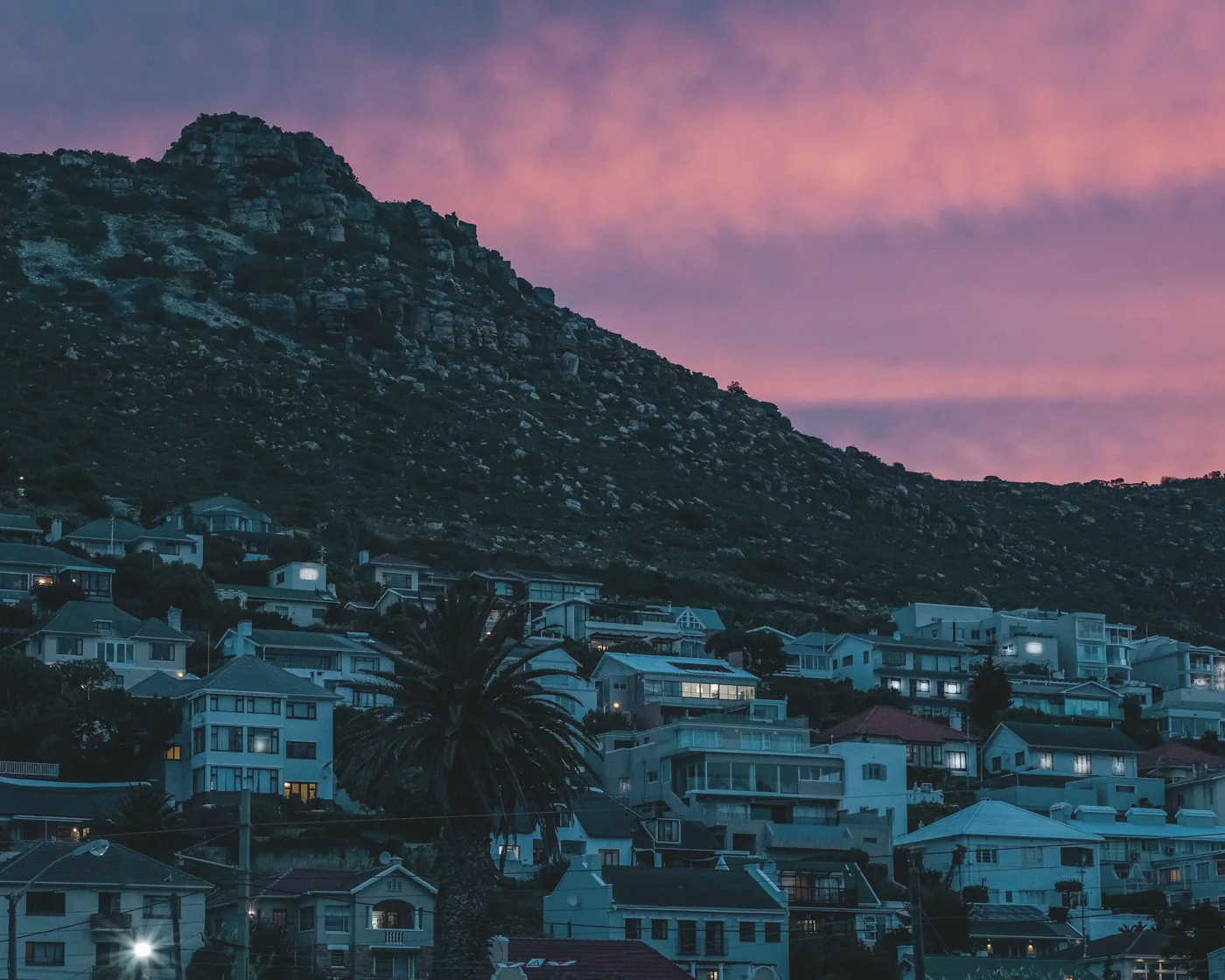 Houses on a hillside above Fish Hoek lighting up at dusk, with a mountain behind and a pink evening sky