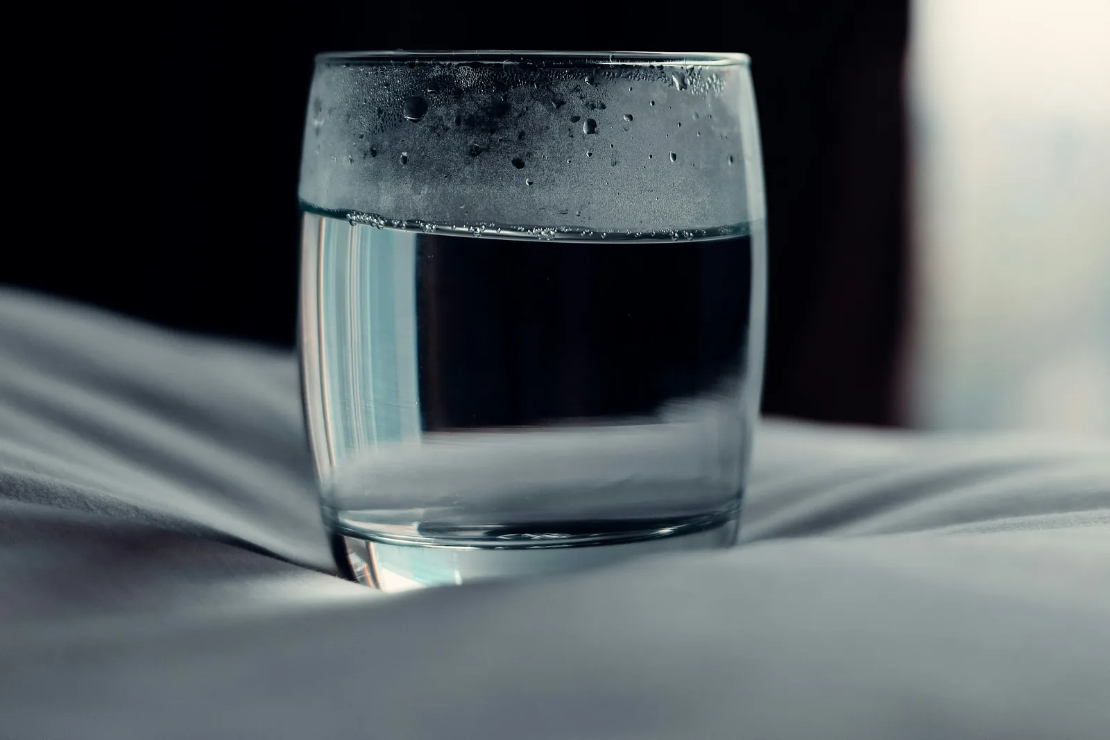 A glass of water on a rumpled bed at night, condensation on the rim, a dark curtain behind it
