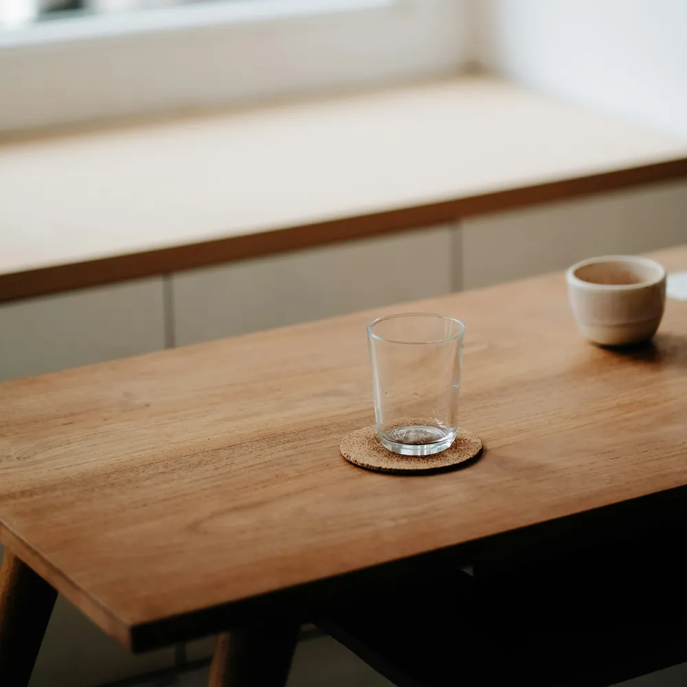 An empty glass tumbler on a cork coaster on a wooden table in soft morning light