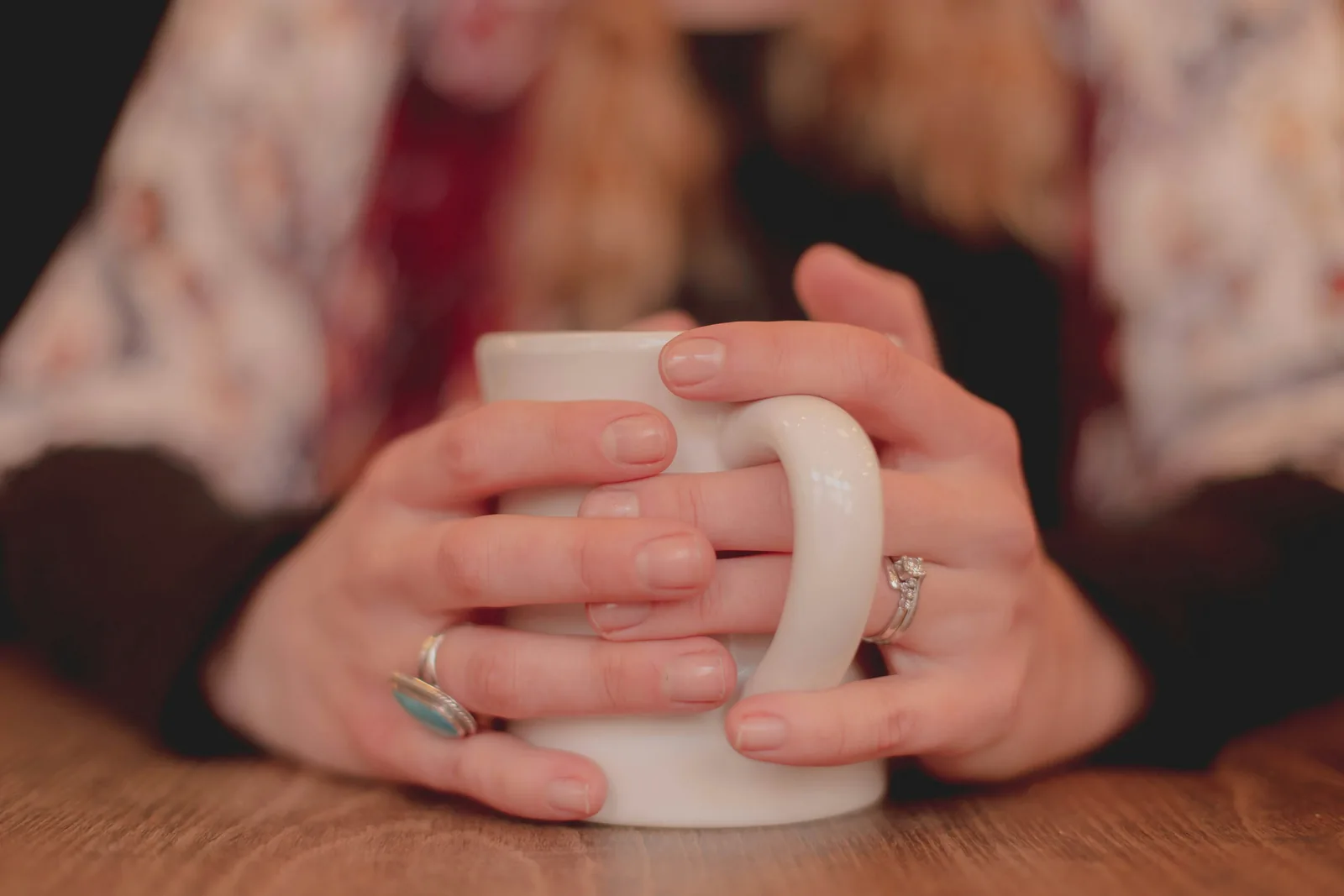 Two hands wrapped around a plain white mug on a wooden table, rings visible, a warm blurred room behind