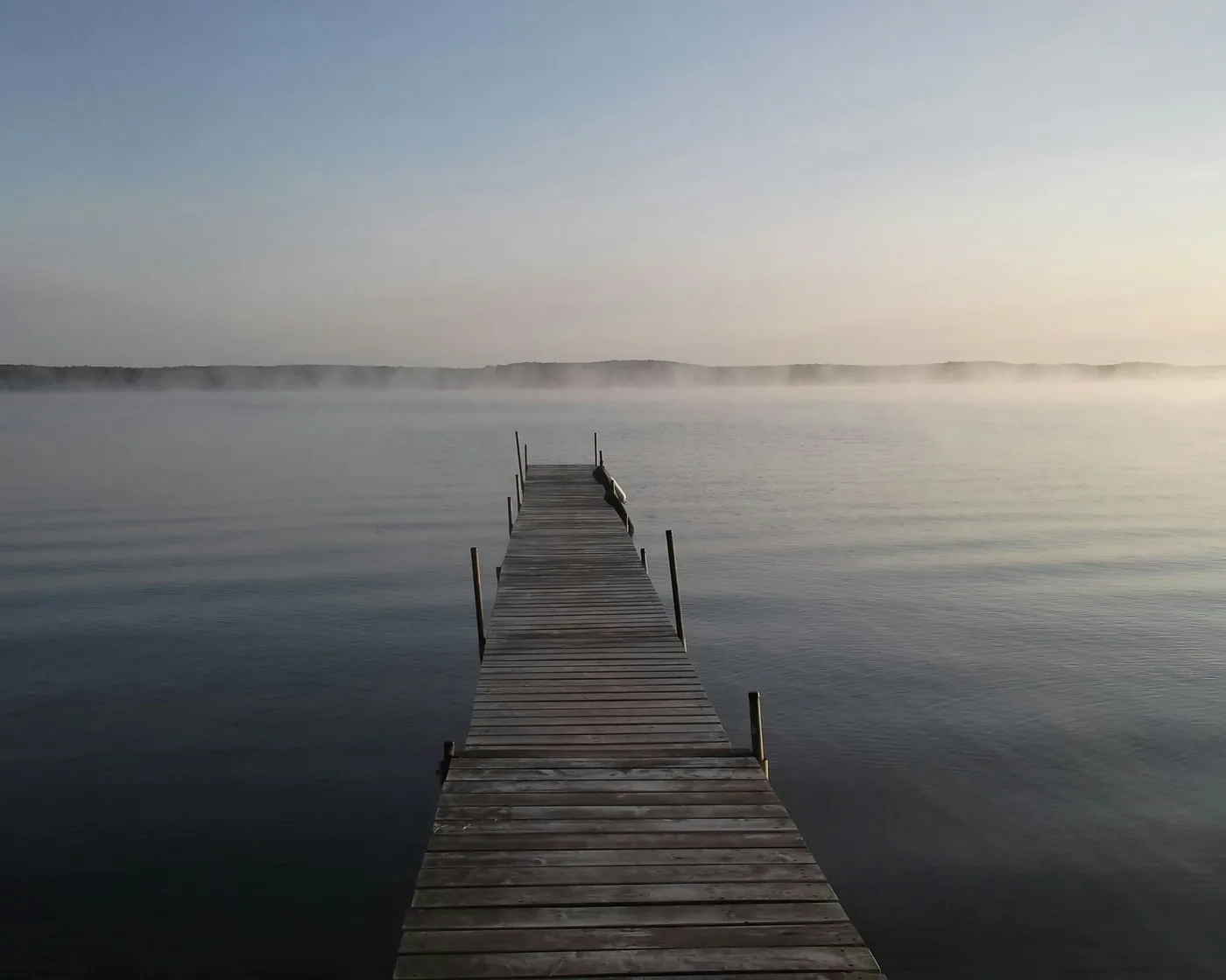 A wooden pier reaching out into still, misty water at dawn, a thin band of low fog along the far shore