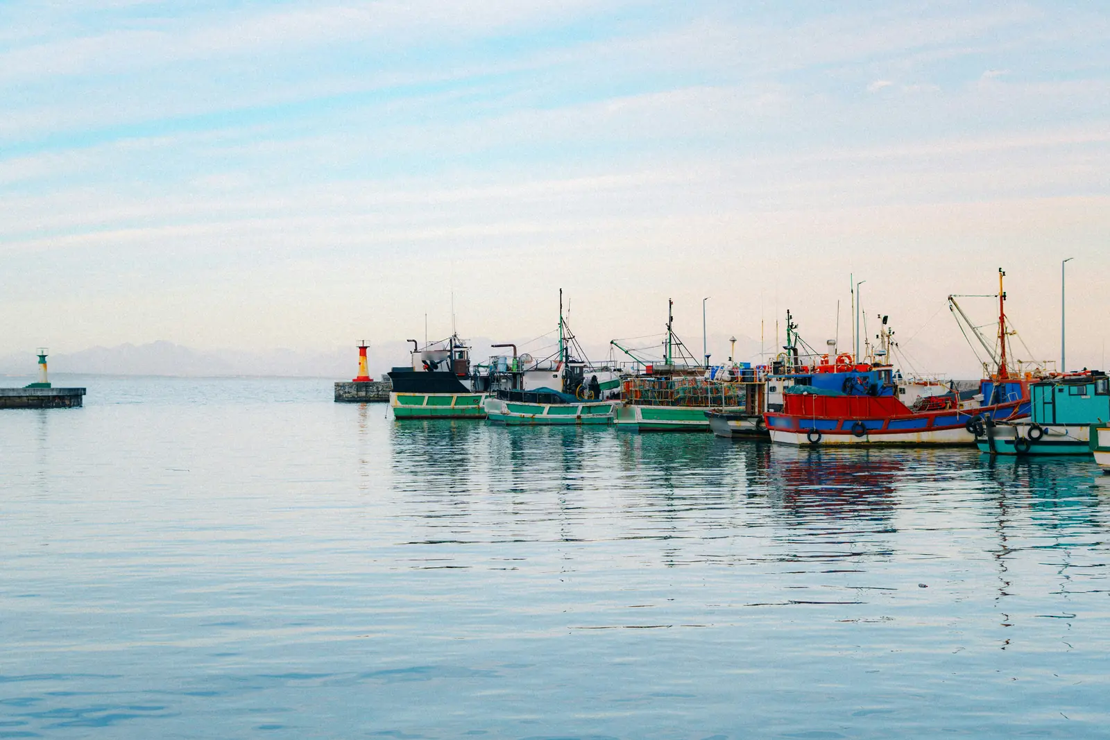 Fishing boats moored on calm water in Kalk Bay harbour, Cape Town, with a small red harbour light and a pale sky