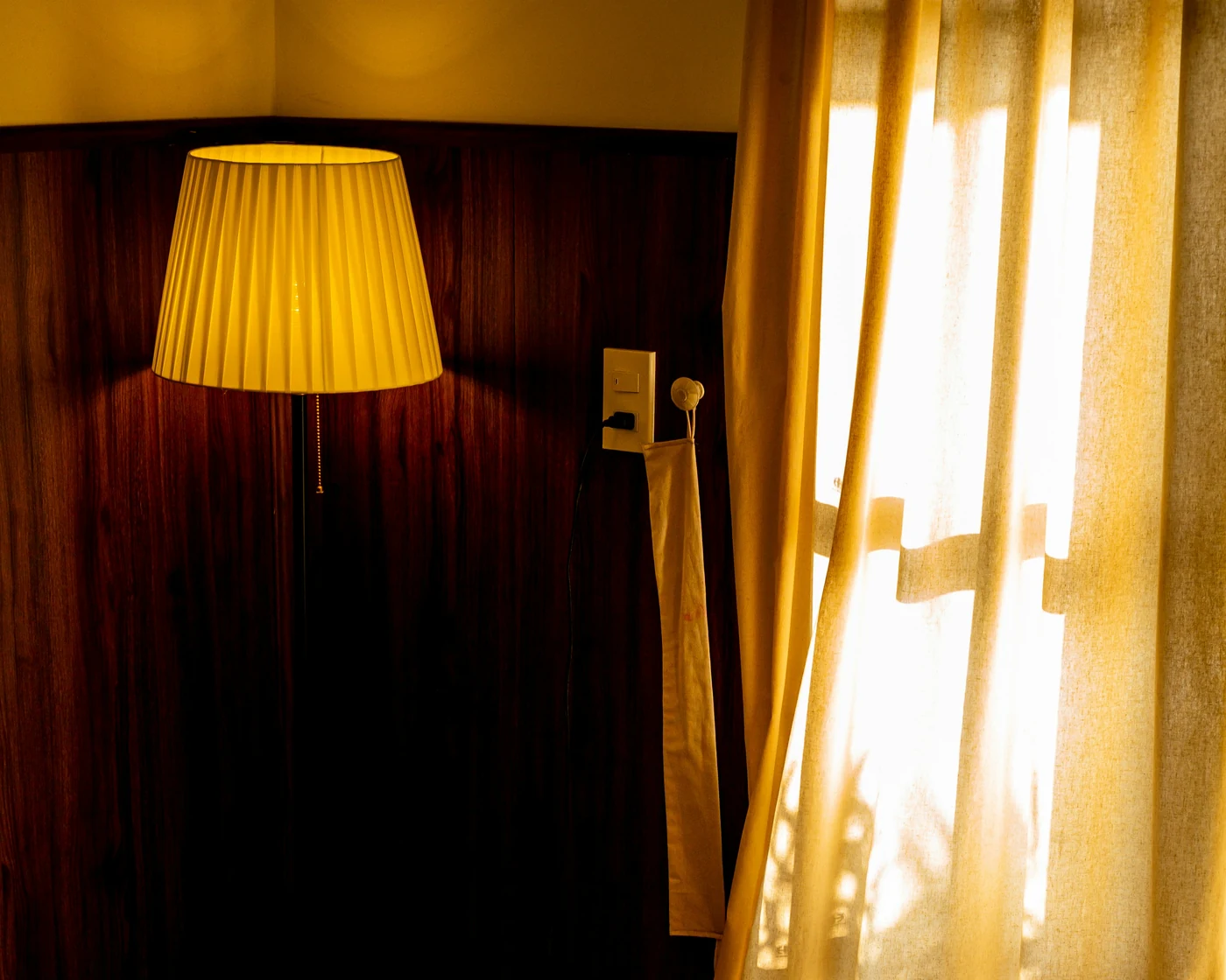 A pleated lamp glowing beside sheer curtains at dusk