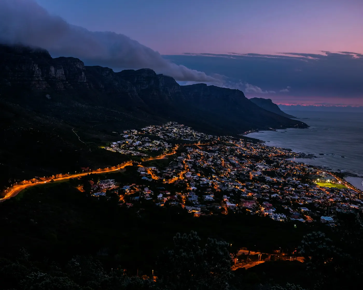 Aerial view of a coastal suburb lit up at night below Lion's Head, with a pink dusk sky and cloud spilling over the mountain, Cape Town