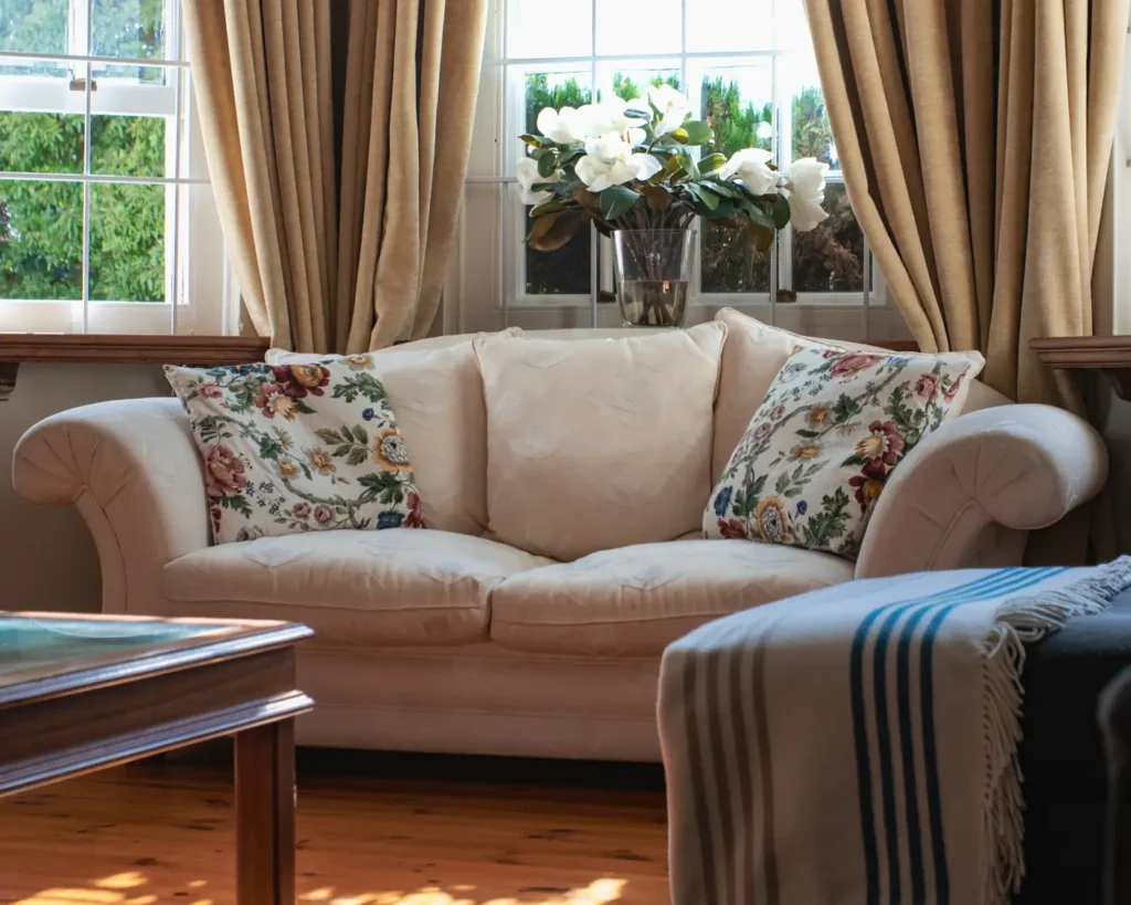 A cream two-seater sofa with floral cushions in a sunlit bay window, with white magnolias and a striped throw