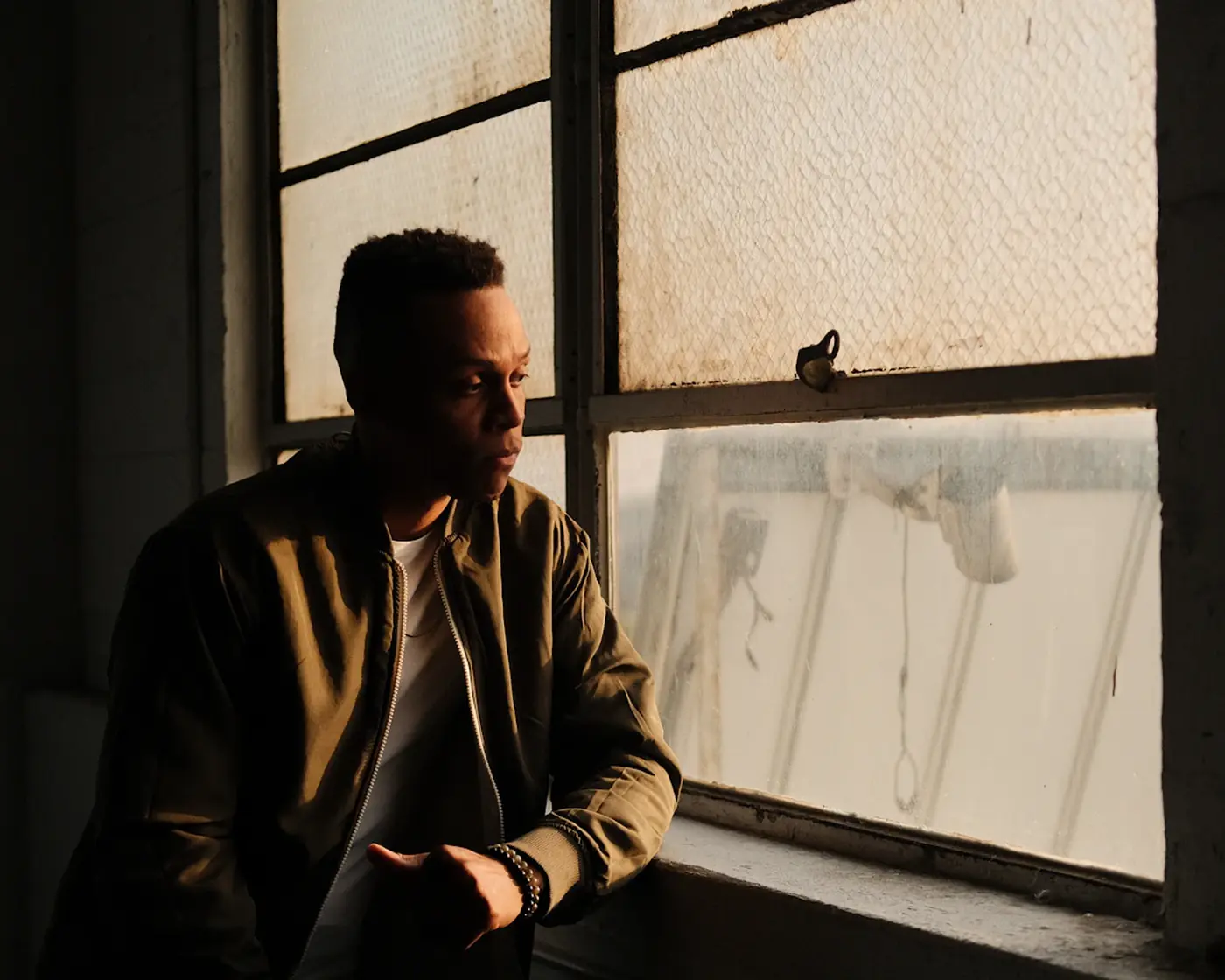 A man in an olive jacket sits by a large factory window, looking down in thought in warm evening light