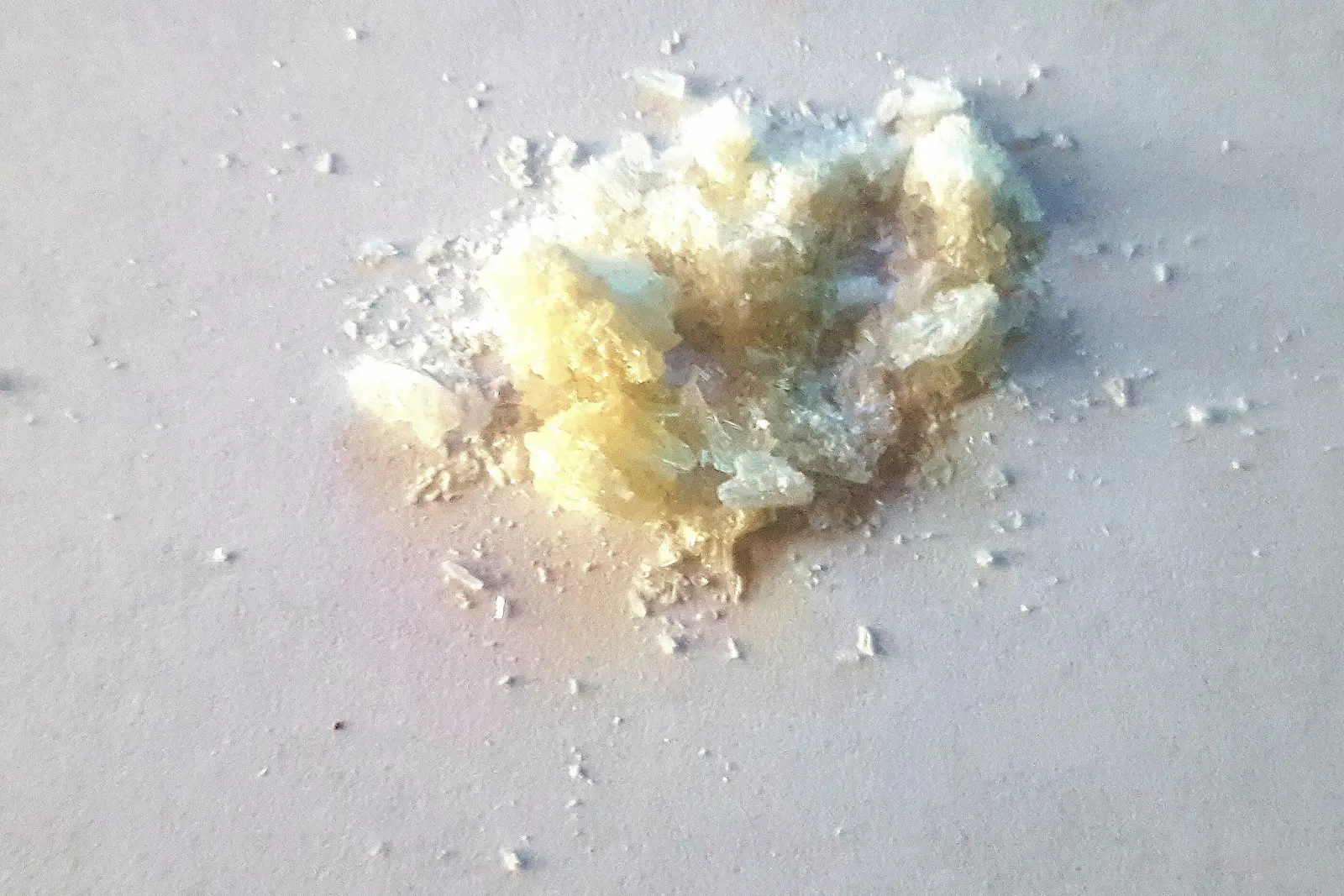 Macro shot of a small pile of white and yellowish mephedrone crystals on a plain light surface, no packaging or text visible