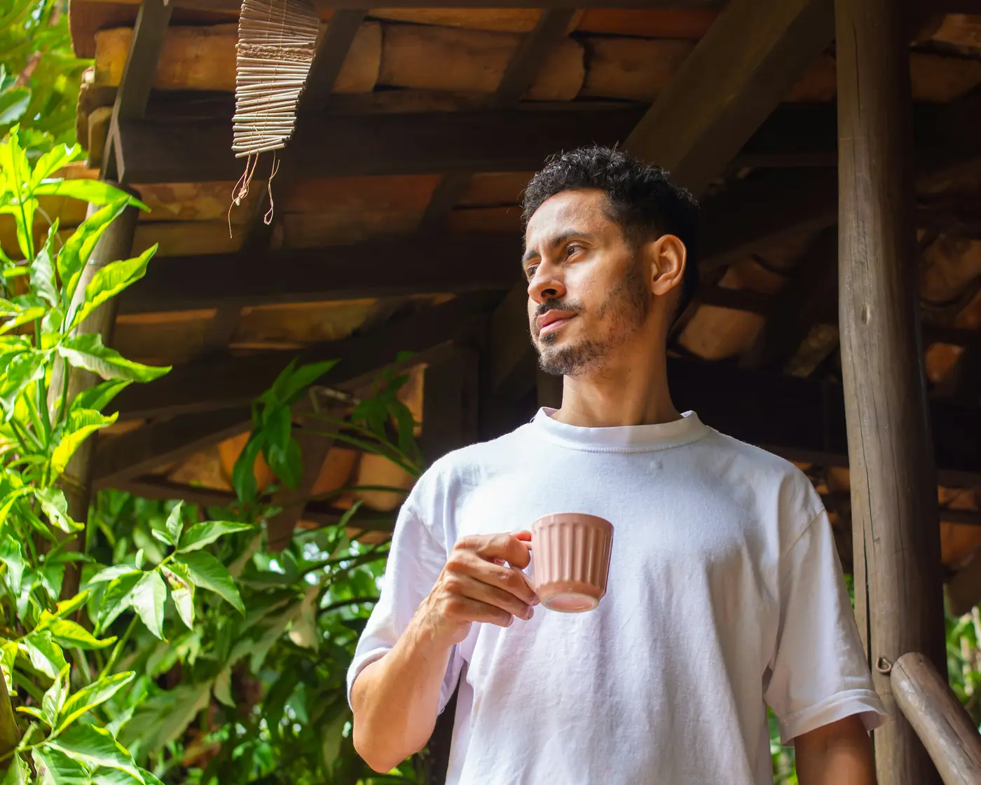 A man in a white T-shirt holds a pink coffee cup on a wooden veranda framed by green leaves in morning light