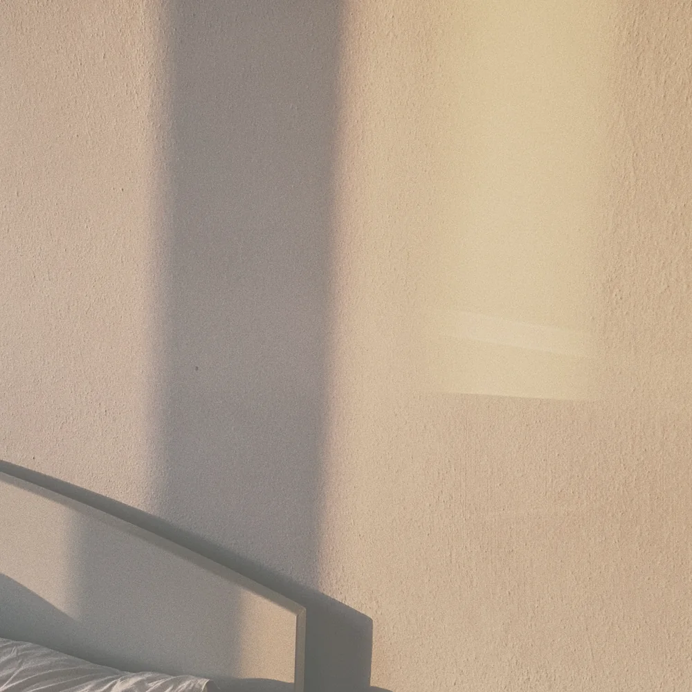 Morning light and a shadow falling across a plain bedroom wall, with the corner of a headboard and pillow below