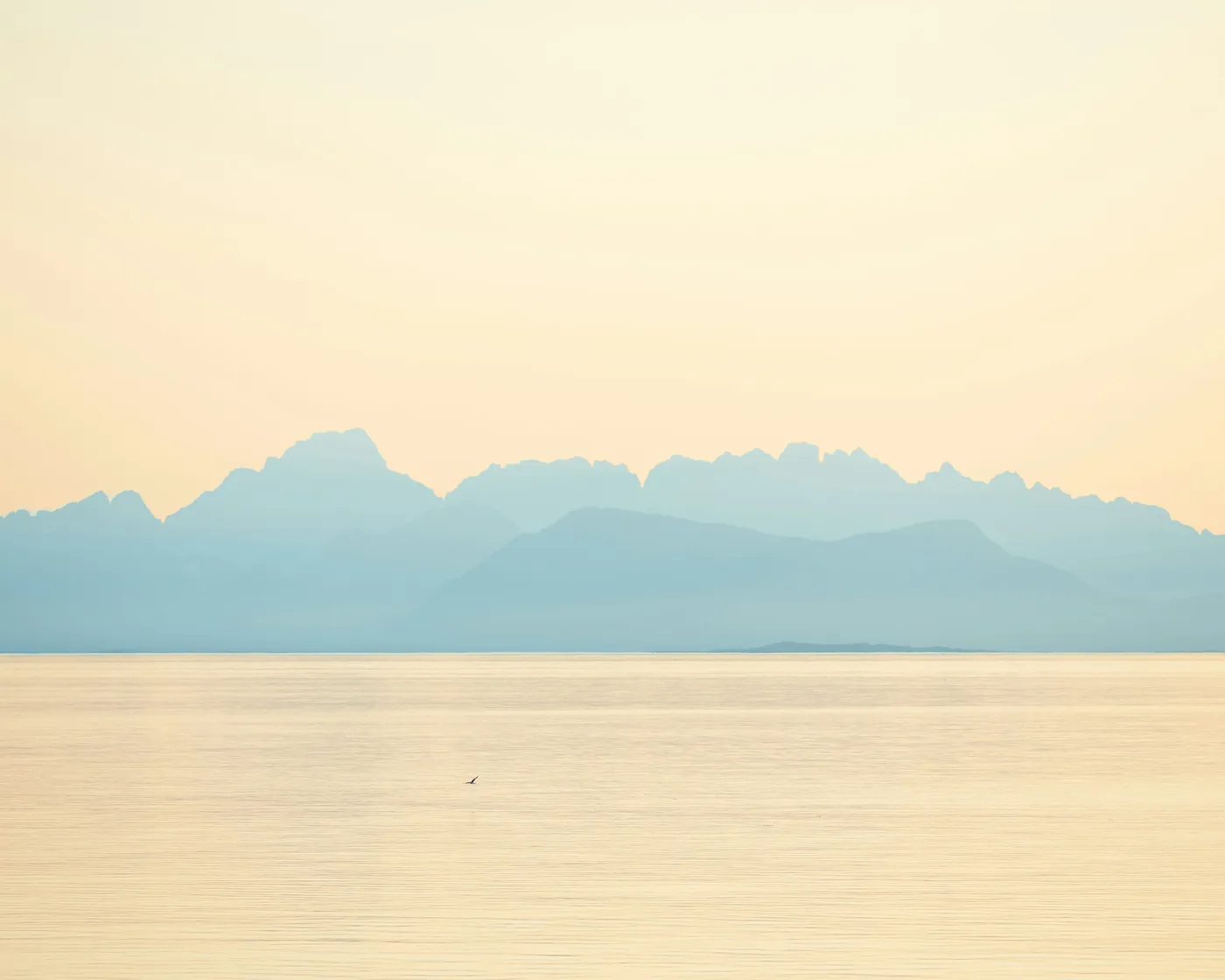 Pale layered mountains fading into mist across a calm sea at dusk, the water reflecting soft gold and blue light