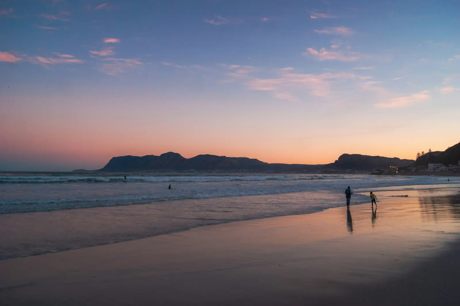 Two people walking along Muizenberg beach in Cape Town at dusk, with mountains under a pink evening sky