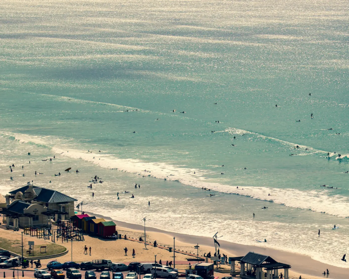 Surfers and swimmers off Muizenberg beach in Cape Town, with the beach pavilion and colourful huts on the sand