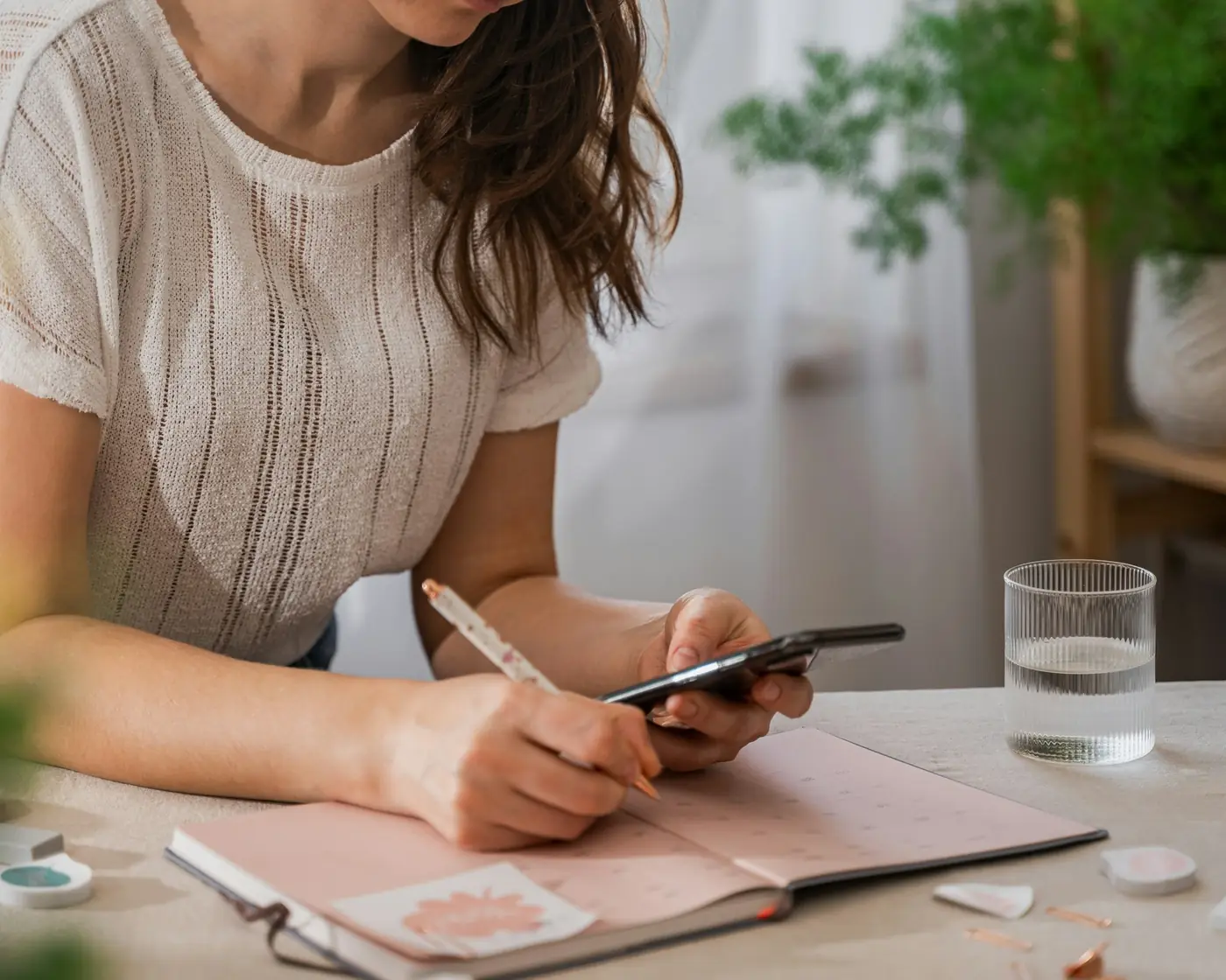 Hands writing notes while checking a phone at a bright kitchen table
