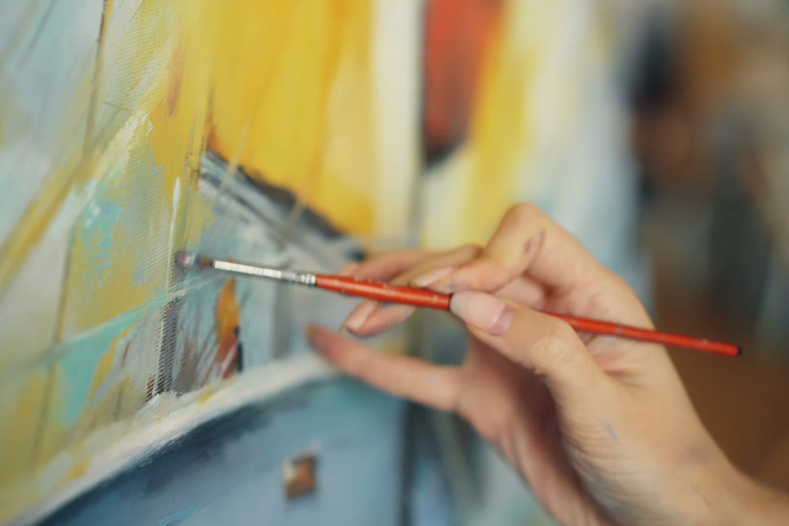 Close-up of a hand holding a red-handled brush, adding a stroke of colour to a warm yellow and blue abstract painting