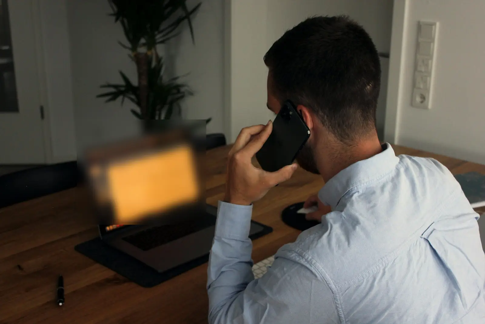 A man on a phone call at a wooden desk, laptop open in front of him