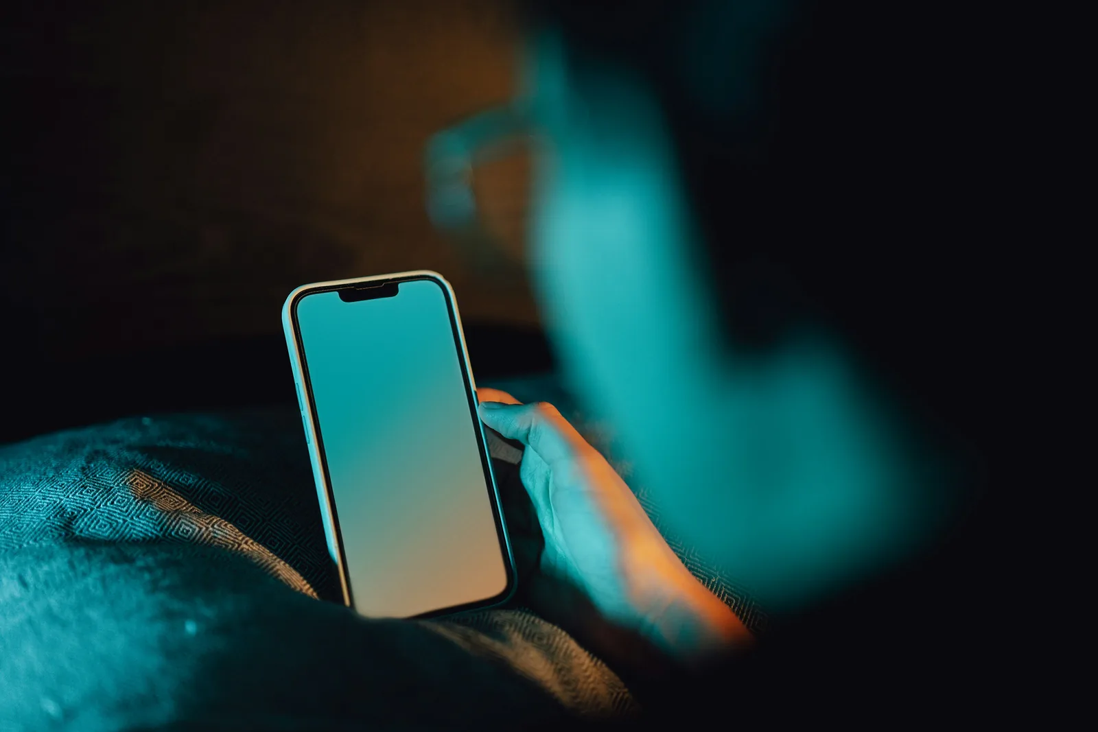 A hand holding a smartphone glowing softly in a dark bedroom