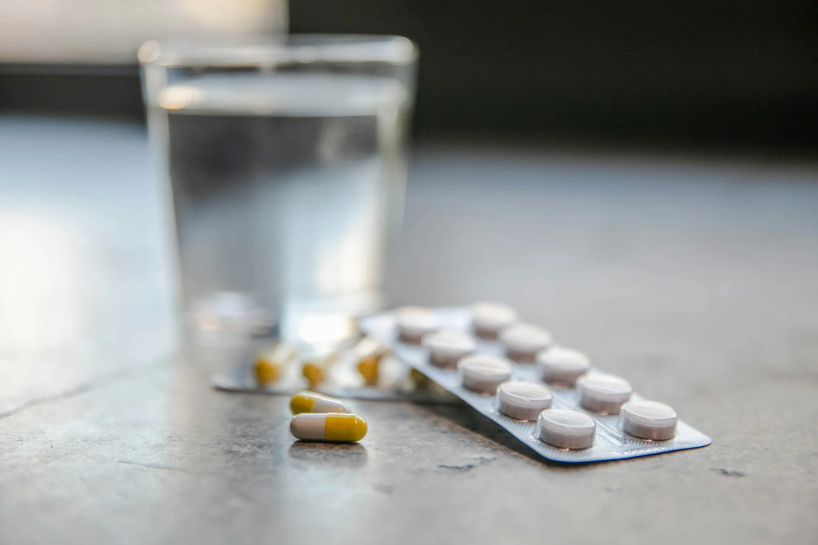 A blister pack of tablets and loose capsules beside a glass of water on a table at night