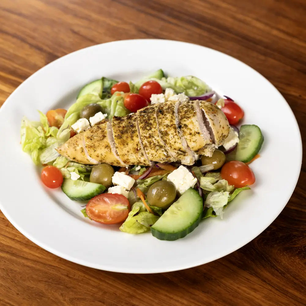 A white plate of grilled chicken salad with feta, olives, cucumber and tomatoes on a dark wooden table