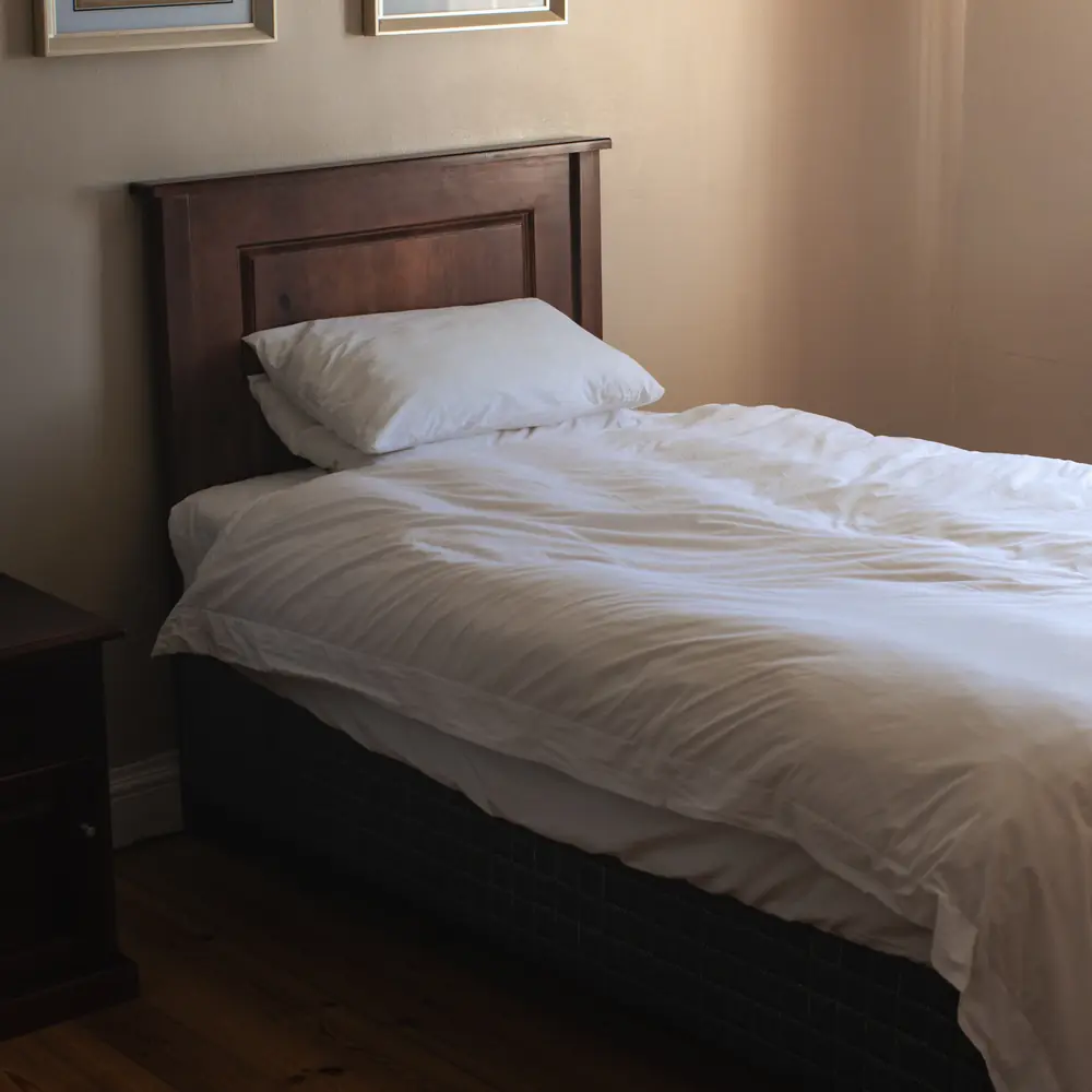 A made single bed with white linen and a dark wooden headboard in a softly lit bedroom with two botanical prints