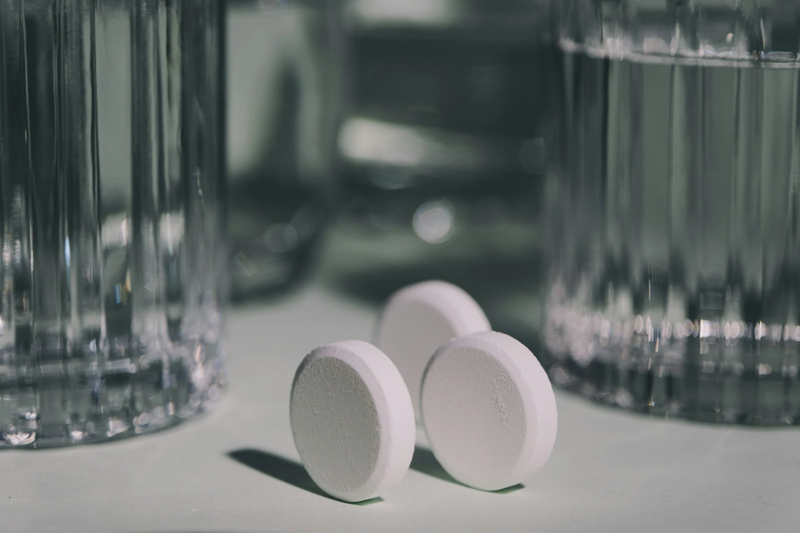 Two white round tablets on a pale table beside blurred glasses of water