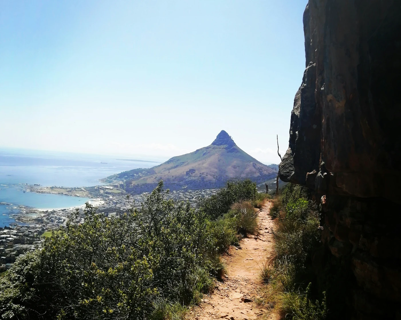 Narrow dirt trail winding along a rocky mountainside towards Lion's Head, with Cape Town and the sea spread out far below